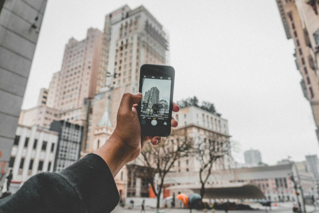 Crop anonymous person taking pictures of modern buildings located on urban street by using mobile phone during autumn cold day