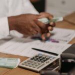 Hands counting euro bills on a wooden desk with calculator, financial documents, and laptop nearby.