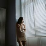 A woman in soft morning light stands by the window, holding a cup and saucer, savoring a peaceful moment.
