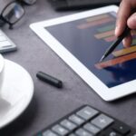 A close-up of a hand using a stylus on a tablet displaying bar graphs, surrounded by office items.