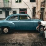 Man cleaning a classic blue car on a gritty urban street, depicting a vintage and rustic setting.