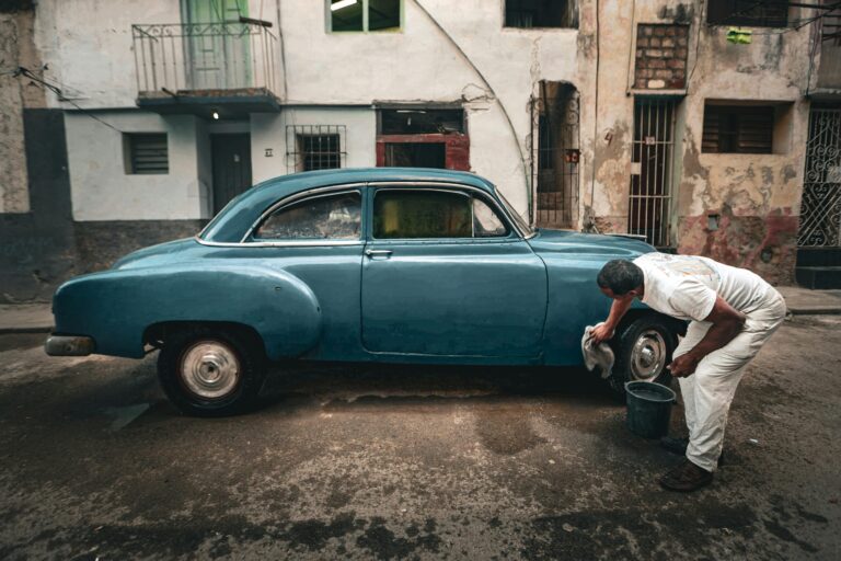 Man cleaning a classic blue car on a gritty urban street, depicting a vintage and rustic setting.