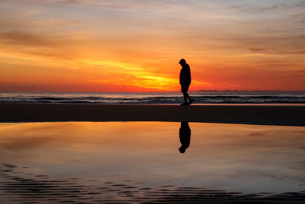 A silhouette of a man walking along the beach at sunrise with a reflective ocean surface.