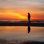 A silhouette of a man walking along the beach at sunrise with a reflective ocean surface.