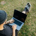 Back view of a young man sitting on grass using a laptop outdoors, showcasing relaxed freelance work lifestyle.