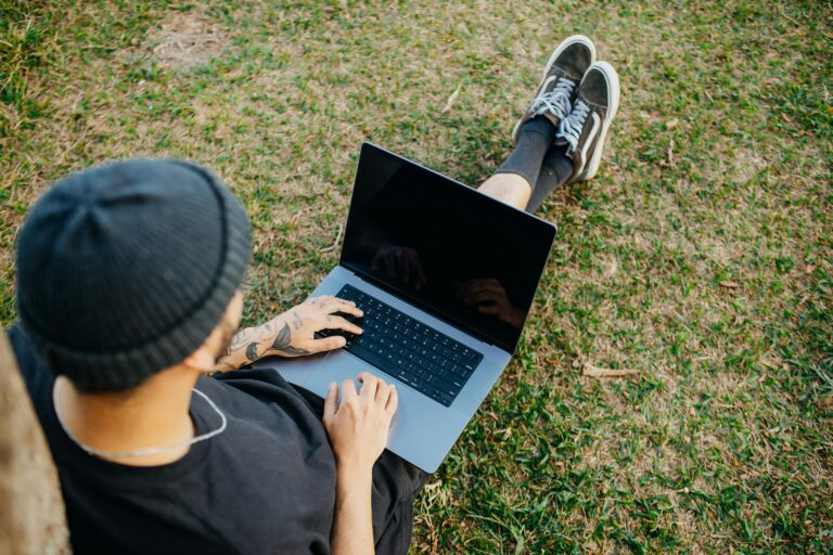 Back view of a young man sitting on grass using a laptop outdoors, showcasing relaxed freelance work lifestyle.