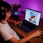 Young girl using a laptop with headphones under purple lighting, focused on photo editing.