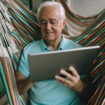Elderly man enjoying leisure time in hammock while using a tablet indoors.