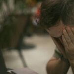 Adult male appears stressed, sitting indoors with his hands on his face beside a laptop.