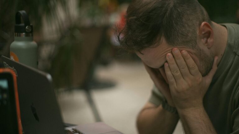 Adult male appears stressed, sitting indoors with his hands on his face beside a laptop.