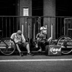 Black and white of anonymous delivery male in face masks sitting near fence and roadway with parked bicycles in town during COVID 19 pandemic