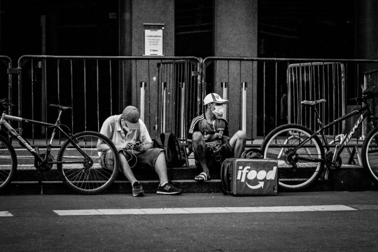 Black and white of anonymous delivery male in face masks sitting near fence and roadway with parked bicycles in town during COVID 19 pandemic
