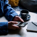 A man uses a smartphone while enjoying coffee, pen, and notepad in a cafe.