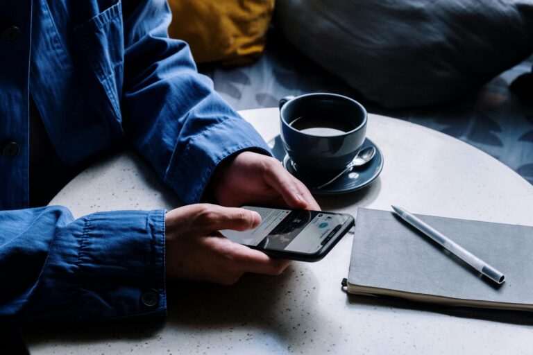 A man uses a smartphone while enjoying coffee, pen, and notepad in a cafe.