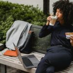 Adult woman sipping coffee and eating a sandwich while working outdoors on a bench.
