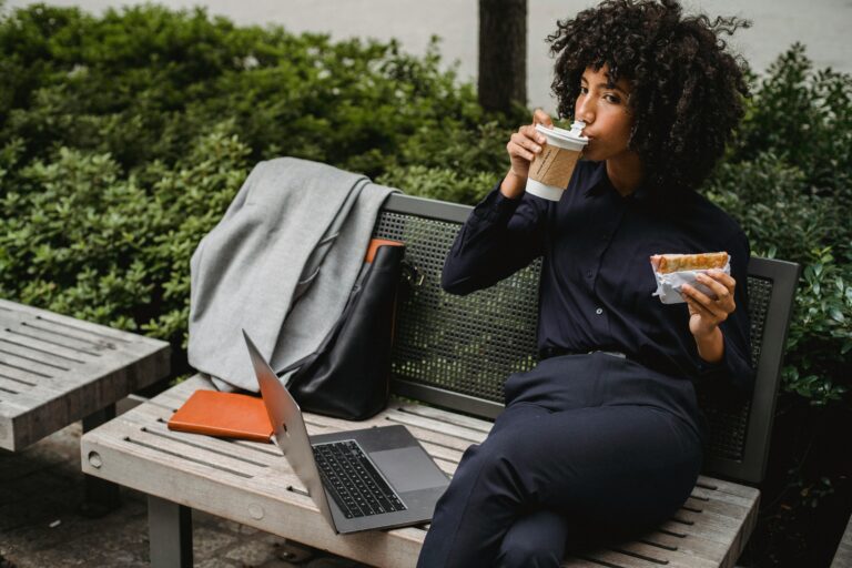 Adult woman sipping coffee and eating a sandwich while working outdoors on a bench.