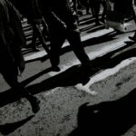 From above black and white of crop anonymous people in warm clothes walking on asphalt roadway with shadows in town