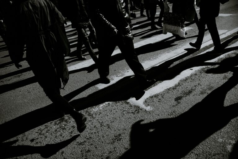 From above black and white of crop anonymous people in warm clothes walking on asphalt roadway with shadows in town