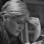 Black and white image of a woman deep in thought, holding a smartphone indoors.