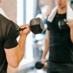 Unrecognizable male athlete with towel pumping biceps with dumbbells while standing near mirror during fitness training in gym on blurred background