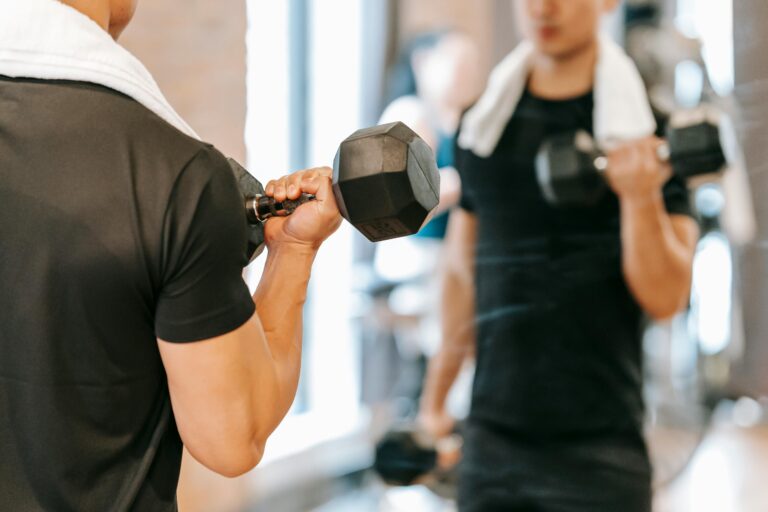 Unrecognizable male athlete with towel pumping biceps with dumbbells while standing near mirror during fitness training in gym on blurred background