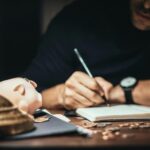 Crop man sitting at desk with vintage lamp and scattered coins and piggybank while writing in notepad
