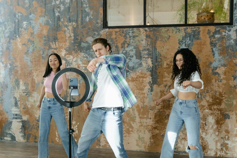 Group of friends dancing indoors with a smartphone and ring light setup for social media content.