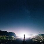 A silhouette of a person standing on a road under a vast, starry night sky with distant light.