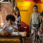 Film crew capturing a scene in a lively cafe setting with diverse actors and a cameraman.