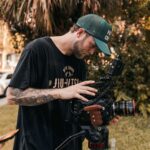 A young male cinematographer adjusts a cinema camera outdoors in a sunny setting.