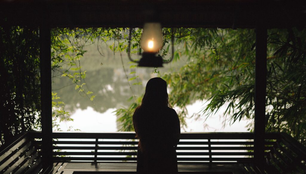 Silhouette of a woman seated on a bench, gazing at a tranquil lake surrounded by lush greenery.