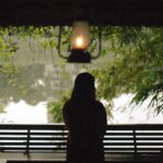 Silhouette of a woman seated on a bench, gazing at a tranquil lake surrounded by lush greenery.