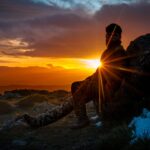 A person sits on a mountain rock at sunset, creating a stunning silhouette against the vibrant sky.