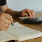 Close-up of hands writing calculations in a notebook with a calculator, focused on budgeting or financial work.