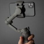 Hand holding a smartphone mounted on a stabilizer against a solid background with tattooed fingers.