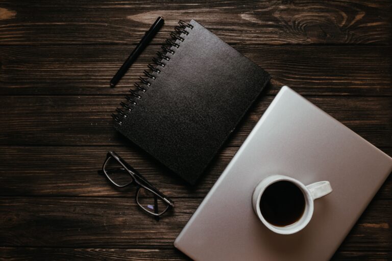 Top view of a wooden desk with a cup of coffee, notebook, pen, and laptop, perfect for productivity.