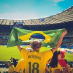 Excited Brazilian fans holding flag at soccer match in vibrant stadium atmosphere.