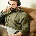Tattooed man in hoodie with headphones working on laptop from home, embodying modern remote work lifestyle.