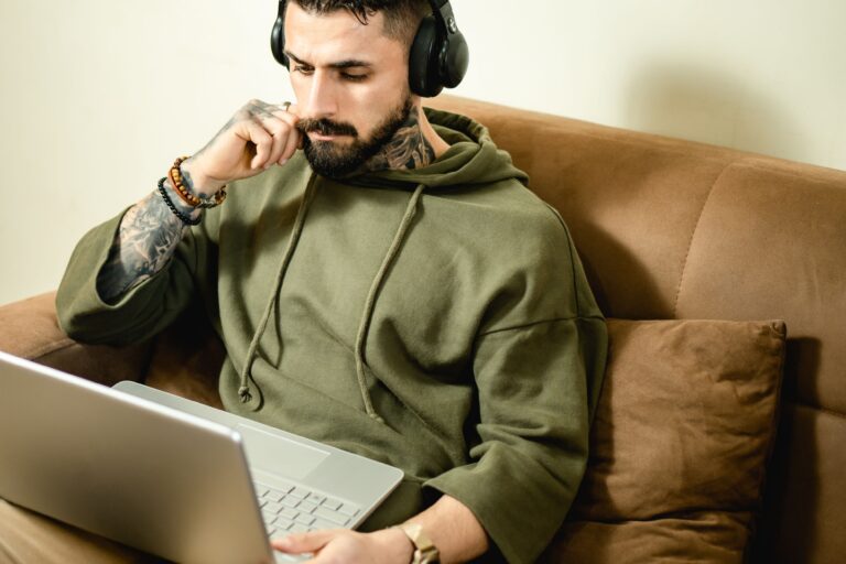 Tattooed man in hoodie with headphones working on laptop from home, embodying modern remote work lifestyle.